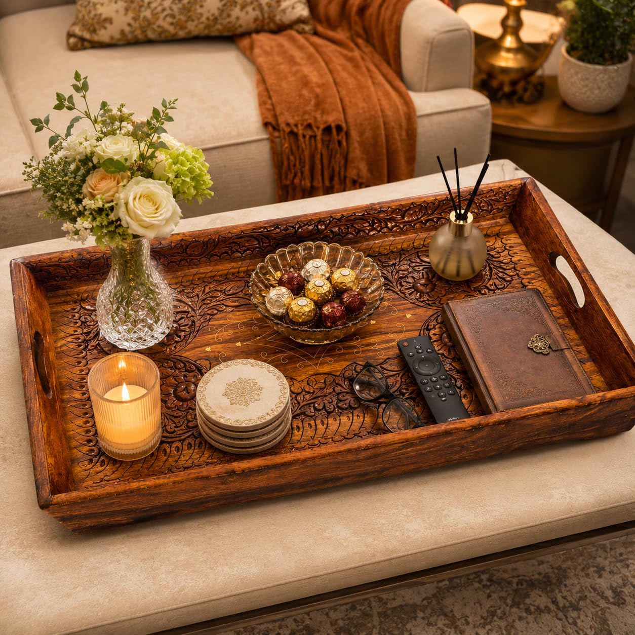 An ornate carved wooden serving tray with cutout handles, styled on a beige sofa in a living room setting. The tray contains a glass vase with white and green flowers, a lit candle, a bowl of chocolates, coasters, eyeglasses, a remote, and a small leather notebook.