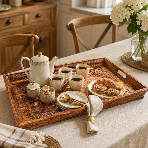 A styled breakfast scene on a dining table featuring a carved dark brown wooden serving tray holding a white ceramic teapot, cups, sugar bowl, small plate, and pastries, with a beige tablecloth and wooden chairs in the background.